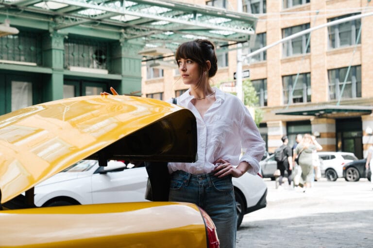 Pessoa ao lado de um carro amarelo com o porta-malas aberto, vestindo jeans e camisa branca, em um cenário urbano movimentado com prédios e transeuntes ao fundo.