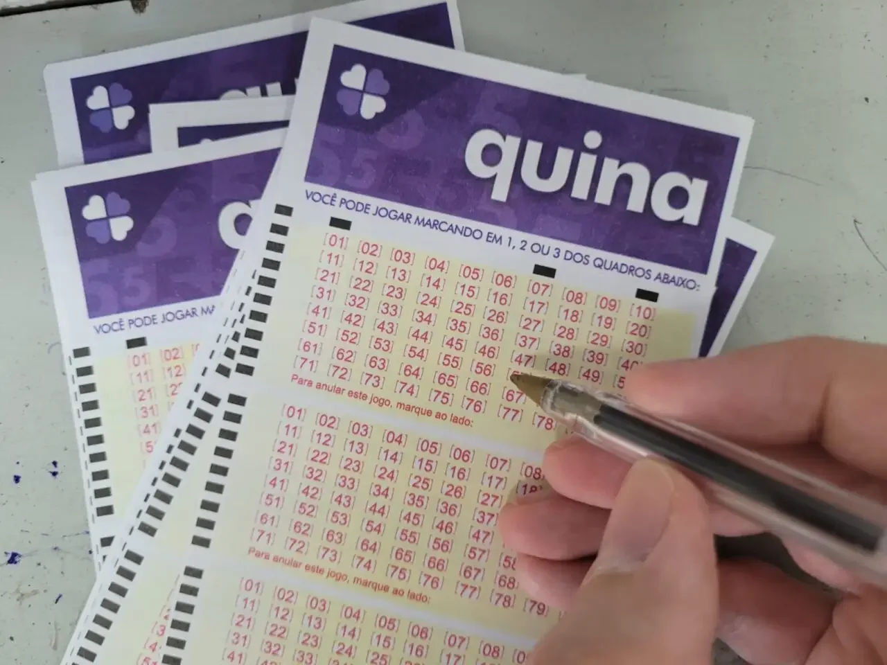 Quina acumula novamente e prêmio chega a R$ 10 milhões: veja os números sorteados 5 Pessoa marca número em bilhete da Quina, com grades numeradas de 01 a 80 e cabeçalho roxo com o nome da loteria brasileira.