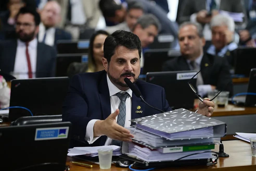 Renato Rodrigues participa de uma sessão formal em um ambiente institucional, rodeado de documentos e equipamentos de trabalho, indicando envolvimento ativo em discussões políticas.