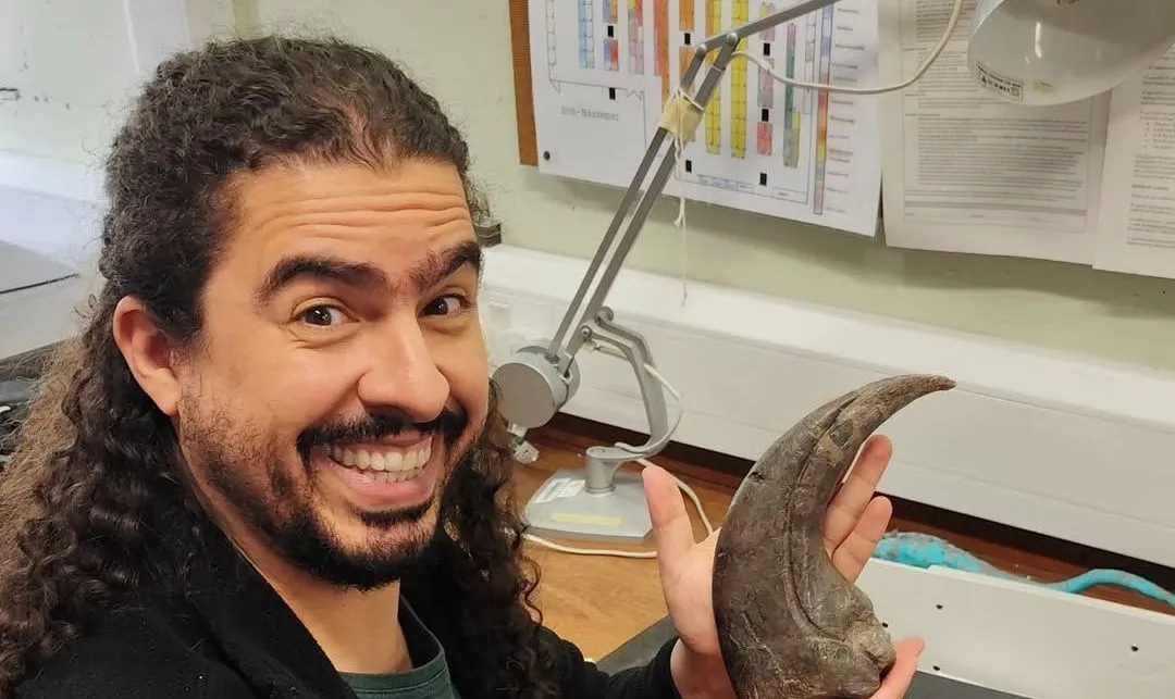 Pessoa segurando uma garra fóssil curvada de dinossauro em ambiente de estudo, com papéis científicos e luminária ao fundo.