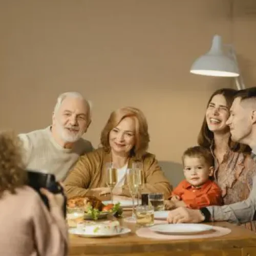 Família sorridente posa durante refeição festiva enquanto alguém registra o momento com uma foto.