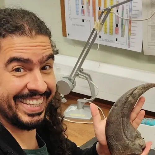Após receber alta hospitalar, o biólogo e divulgador científico Pirulla retorna aos estudos com garra — literalmente. A imagem mostra o youtuber em seu ambiente de pesquisa, segurando uma impressionante garra fóssil.